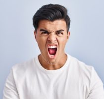 Hispanic,Man,Standing,Over,Blue,Background,Angry,And,Mad,Raising